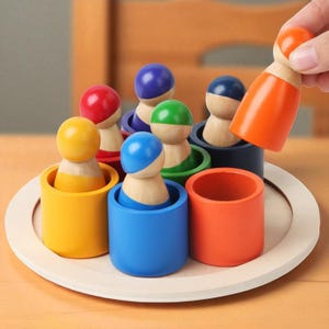Puede incluir: Un juguete educativo de madera con tazas cilíndricas de colores y muñecos de clavija a juego. Las tazas son de color amarillo, azul, verde, rojo y naranja. Un muñeco de clavija naranja se sostiene sobre una taza. El juguete está sobre una bandeja de madera.