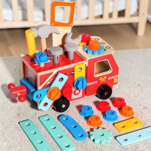 Può includere: Un camion dei pompieri giocattolo in legno con vari strumenti e accessori colorati. Il camion è rosso con ruote nere e presenta un martello, una chiave inglese e viti. Sono inclusi pezzi di legno blu, verdi e gialli.