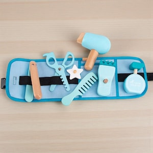 Pode incluir: Um organizador de tecido azul contém brinquedos de cabeleireiro de madeira. O conjunto inclui um secador de cabelo azul claro, tesoura, pente e um borrifador. Também inclui um modelador de cabelo de madeira e um acessório em forma de estrela.
