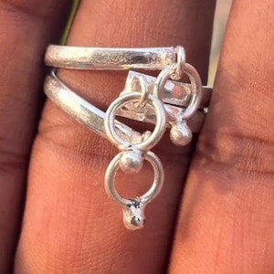Puede incluir: Un anillo de plata con un diseño único. El anillo presenta dos bandas, una encima de la otra, con colgantes circulares y pequeños detalles de bolas. El anillo se lleva en un dedo, mostrando sus intrincados detalles y artesanía.