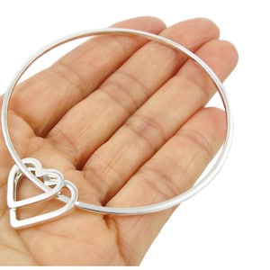 May include: A silver bangle bracelet featuring two intertwined heart charms. The bracelet has a simple, elegant design with a polished finish. The hearts are connected to the main band of the bracelet, creating a delicate and romantic look.