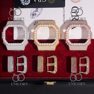 May include: Three bejeweled watch cases and matching buckles in silver, gold, and rose gold. Each case is encrusted with small, sparkling stones. The buckles are also adorned with similar stones. The items are displayed on a red velvet surface.