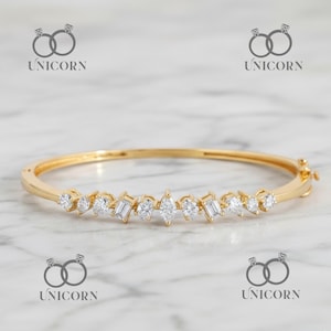 Op de afbeelding: Een gouden armband met heldere edelstenen in verschillende vormen. De armband heeft ronde, baguette en peervormige stenen. De sluiting is rechts zichtbaar. De achtergrond heeft een licht marmerpatroon. Een sieraad.