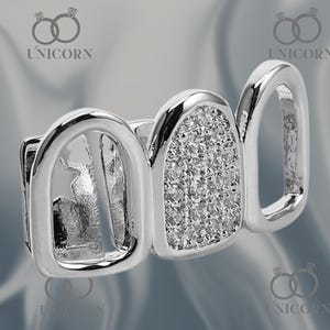 May include: Silver-toned grill with three teeth visible. The center tooth is encrusted with small, sparkling stones. The grill has a polished finish and is designed to fit over teeth. The brand name "UNICORN" is visible.