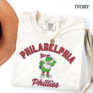 May include: An ivory-coloured t-shirt with the word "PHILADELPHIA" in red arched lettering above a cartoon mascot. The mascot is green with a red and white striped shirt and is holding a flag. The word "Phillies" is written in red script below.