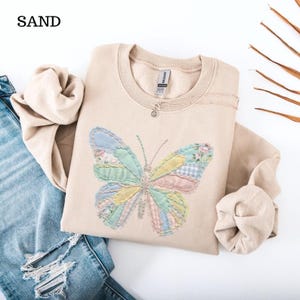 Può includere: Una felpa color sabbia con un design a farfalla patchwork nei colori pastello. La farfalla è realizzata con vari modelli di tessuto, tra cui floreale e gingham. È visibile anche una collana d'argento con un piccolo ciondolo.