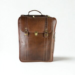 May include: A brown leather briefcase with a crocodile skin texture. The bag has a top handle, a flap closure with two buckles, and a brass clasp. The stitching is white, and the bag has a zipper on the side.