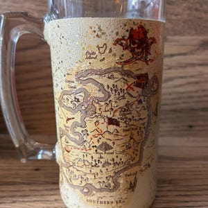 May include: A clear glass beer mug featuring a textured, beige-colored wrap with a printed treasure map. The map includes illustrations of a skull and crossbones, Maya Ruins, and text labels such as Pirate Island, Scorpion Islands, and Southern Sea.