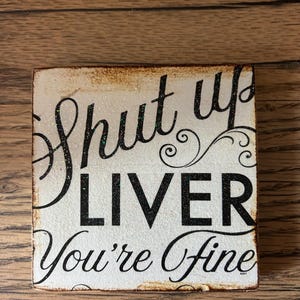 Puede incluir: Un letrero cuadrado de madera envejecida con las palabras "Shut up LIVER You're Fine" en letras negras. El letrero tiene un fondo blanco con un borde brillante y está sobre una superficie de madera.