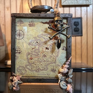 Peut inclure: Une lanterne en bois avec un motif de carte de pirate. La carte est de couleur beige avec des détails bruns et présente une rose des vents, des navires et le texte "Southern Sea". Un charme de tête de mort et d'os croisés est suspendu en haut. Des coquillages ornent les coins et une poignée en corde complète le design.