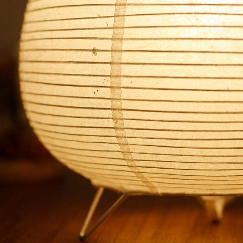 Puede incluir: Una l&aacute;mpara de papel esf&eacute;rica que emite una luz c&aacute;lida y suave. La pantalla blanquecina presenta l&iacute;neas horizontales y una costura vertical. La l&aacute;mpara se apoya en tres finas patas de color plateado, creando un dise&ntilde;o minimalista.