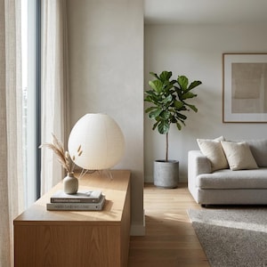 Puede incluir: Un sal&oacute;n moderno con una l&aacute;mpara de papel redonda y blanca sobre un mueble de madera. Un peque&ntilde;o jarr&oacute;n con plantas secas est&aacute; junto a la l&aacute;mpara. Tambi&eacute;n se ve una planta verde en maceta y un sof&aacute; gris claro con cojines.