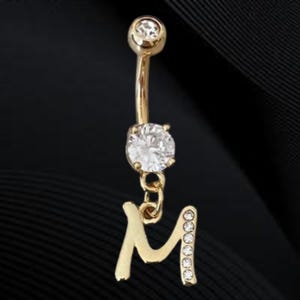 May include: Gold-toned belly button ring with a curved barbell design. The top and bottom feature clear, round gemstones. A dangling letter "M" is attached, embellished with smaller, clear gemstones.