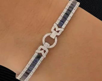 May include: A silver bracelet with a geometric design. The bracelet features alternating rows of small, square, dark blue stones and clear, round stones. The clasp is a circular design with clear stones.