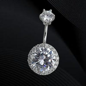 Op de afbeelding: Een zilveren navelpiercing met een grote, ronde, heldere edelsteen omringd door kleinere stenen. De bovenste steen is een kleinere, stervormige heldere edelsteen. De sieraden zijn geplaatst tegen een donkere achtergrond.