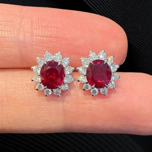 Puede incluir: Un par de pendientes de botón con piedras preciosas rojas oscuras de corte ovalado, cada una rodeada por un halo de piedras preciosas claras más pequeñas. Los pendientes están engastados en un metal plateado, creando un diseño brillante. Las piedras preciosas miden aproximadamente 1,3 cm de largo.