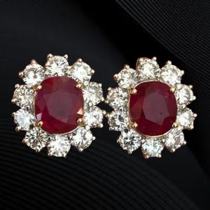 May include: A pair of gold-toned stud earrings. Each earring features a large, oval, deep red ruby surrounded by a halo of smaller, clear, round-cut diamonds. The earrings are set against a black background.