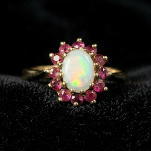 May include: A gold ring featuring a central oval-shaped iridescent opal gemstone surrounded by a halo of twelve small, round, deep pink rubies. The metal band is polished gold, and the stones are set in a classic cluster arrangement.