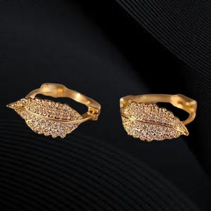 Peut inclure: Une paire de boucles d'oreilles dorées en forme de feuille. Chaque boucle d'oreille présente un motif de feuille détaillé incrusté de petits cristaux étincelants. Les boucles d'oreilles sont posées sur un fond sombre.