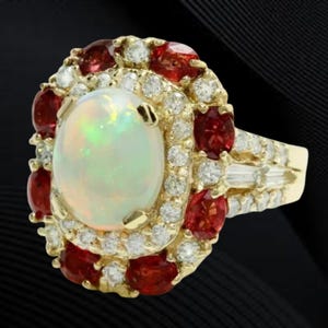 Puede incluir: Un anillo de oro con una gran piedra central de ópalo ovalada, rodeada de pequeños diamantes y piedras rojas. El anillo tiene un diseño de halo con una caña dividida y diamantes adicionales en la banda.