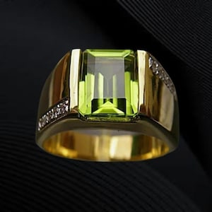 May include: A gold ring featuring a large, rectangular, light green gemstone. The ring has a wide band and is accented with small, clear gemstones on the sides. The ring is set against a dark background.