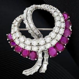 May include: A dazzling brooch featuring a swirling design of white diamonds and vibrant, oval-cut rubies. The brooch has a pin backing and is set in a silver metal. The rubies are a deep, rich red color.