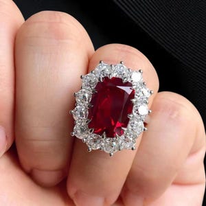Könnte beinhalten: Nahaufnahme eines Rings mit einem großen, rechteckigen, tiefroten Edelstein, umgeben von kleineren, klaren Edelsteinen. Der Ring ist auf einem silbernen Band gefasst, was einen auffälligen Kontrast erzeugt.