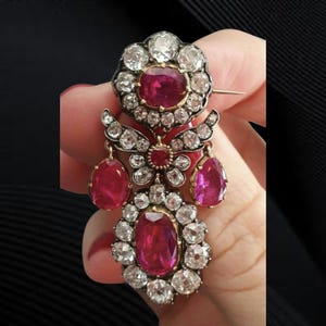 Puede incluir: Broche antiguo con un rubí central ovalado y piedras preciosas claras. El broche tiene un diseño floral con detalles de rubíes y piedras preciosas, y un alfiler dorado.