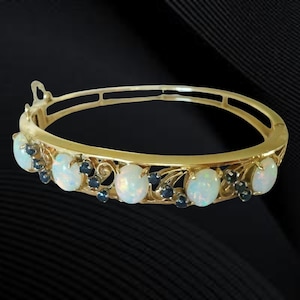 Puede incluir: Un brazalete de oro con ópalos ovalados y pequeñas piedras preciosas azul oscuro. El brazalete tiene un diseño de filigrana decorativa entre los ópalos. El cierre es visible en la parte superior.