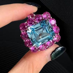 Pode incluir: Um anel ornamentado com uma pedra preciosa quadrada azul clara no centro, rodeada por pedras rosas redondas menores. O design do anel é intrincado e atraente, com foco nas cores vibrantes e no jogo de luz nas pedras preciosas.