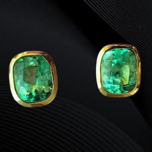 Könnte beinhalten: Ein Paar smaragdgrüne Edelstein-Ohrstecker mit einer goldfarbenen Lünettenfassung. Die Edelsteine haben einen Kissenschliff und sind vor einem dunklen Hintergrund platziert.