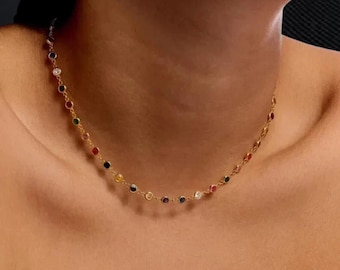 May include: A delicate gold necklace featuring a series of colorful gemstones. The necklace is composed of small, round gemstones in various colors, including red, blue, green, and white, set along a thin gold chain. The necklace is worn around the neck.