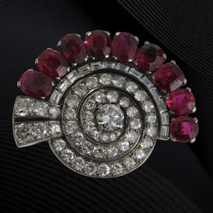 May include: A vintage brooch featuring a spiral design with a central diamond and surrounding smaller diamonds. The brooch is accented with oval-cut rubies. The jewelry is set in a silver or platinum metal.