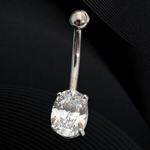 Pode incluir: Um piercing de umbigo prateado com uma grande pedra preciosa clara de corte oval. A barra curva tem uma pequena bola no topo e uma cravação de garras para a pedra preciosa. A joia está sobre um fundo escuro.