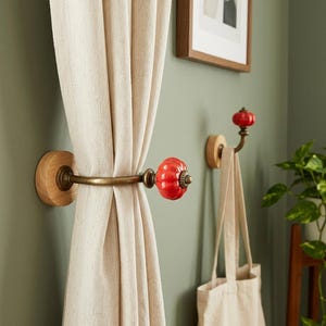 May include: Two wall-mounted hooks featuring a round light wood base, a curved antique brass metal arm, and a glossy red pumpkin-shaped ceramic knob. One hook holds a beige fabric curtain, while the other supports a beige canvas tote bag.