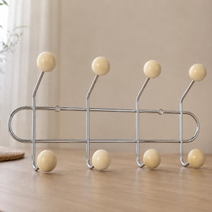 May include: A wall-mounted coat rack featuring a polished chrome metal frame with eight hooks. Each hook is tipped with a smooth, round, cream-colored ball. The rack has a horizontal rectangular structure with rounded ends and vertical supports.