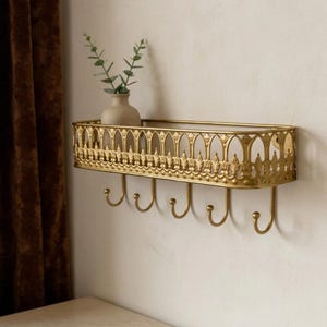 May include: A gold-colored metal wall shelf featuring an ornate, arched lattice railing along the top edge and five curved hooks extending from the bottom. A small, light-colored ceramic vase containing a green plant sits inside the shelf.