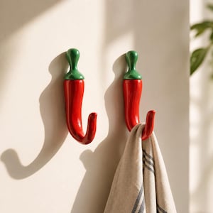 Op de afbeelding: Twee wandhaken in de vorm van rode chilipepers met groene steeltjes. De haken hebben een glanzende afwerking en een gebogen ontwerp. De ene haak is leeg, terwijl de andere een beige stoffen handdoek met dunne zwarte strepen vasthoudt.