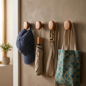 May include: Four rounded light wood wall hooks mounted in a horizontal row. The hooks hold a blue denim baseball cap, a folded beige umbrella, a tan leash with a brass clip, and a small tote bag with a teal and gold fan-shaped pattern.