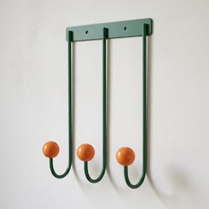 May include: A wall-mounted coat rack featuring a dark green metal frame with three vertical bars. Each bar terminates in a curved hook topped with a smooth, glossy orange sphere. The rectangular mounting plate includes two pre-drilled screw holes.