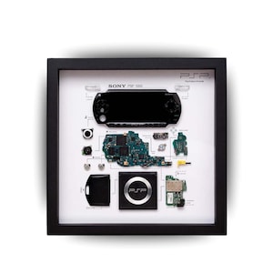Może przedstawiać: Oprawiona, rozłożona na części konsola Sony PSP 1000, prezentująca jej komponenty. Czarna rama otacza białe tło z PSP, płytkami drukowanymi i innymi elementami. Widoczne jest logo PSP. Widoczny jest również tekst "PSP" i "Sony PSP 1000".