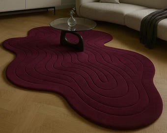 May include: A burgundy-colored, abstract-shaped rug with a wavy, textured design. The rug is placed on a hardwood floor in a living room setting, with a modern sofa, coffee table, and artwork in the background. The rug's unique shape adds a contemporary touch.