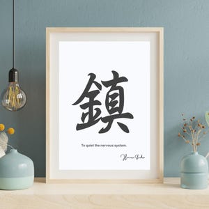 以下が含まれることがあります： 白い背景に黒い漢字が描かれた額入りのアートプリント。文字の下には「To quiet the nervous system」と書かれています。フレームは明るい木材です。