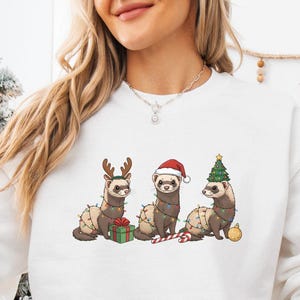 May include: White sweatshirt featuring a festive design of three ferrets adorned with Christmas lights, reindeer antlers, a Santa hat, and a Christmas tree. One ferret holds a gift and another has a candy cane. A Santa mug is held in the hand.