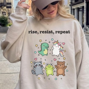 May include: Sand-colored sweatshirt with the text "rise, resist, repeat" above a graphic of a dinosaur, unicorn, raccoon, frog, and bear. The sweatshirt has a crew neck and long sleeves.