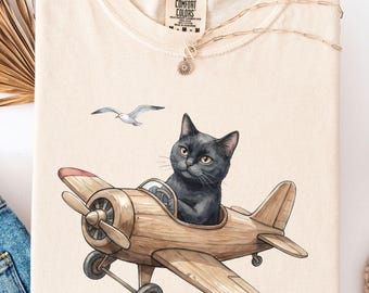 May include: Ivory-colored sweatshirt featuring a black cat piloting a wooden airplane, with a seagull in the sky. The design includes red accents on the plane's wings. The shirt is paired with blue jeans and a necklace.