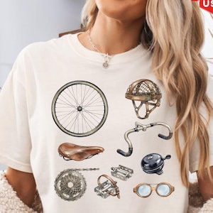 May include: An ivory t-shirt featuring a graphic print of vintage bicycle components, including a wheel, a brown leather saddle, handlebars, a chain, pedals, a bell, a pair of goggles, and a protective leather-style cycling helmet.
