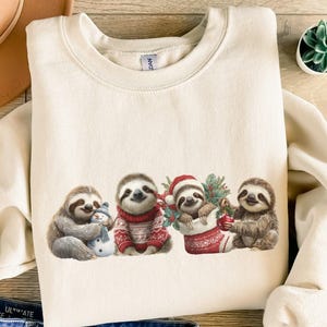 May include: A sand-colored sweatshirt featuring a festive sloth design. Four sloths are depicted, one holding a snowman, another wearing a red sweater, a third in a Christmas stocking, and the last holding a candy cane. The design is printed on a soft, casual sweatshirt.