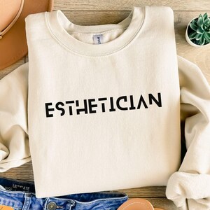 Peut inclure: Un sweat-shirt à col rond de couleur crème avec le mot ESTHETICIAN imprimé en lettres capitales noires sur la poitrine. Le vêtement possède des manches longues et un col, des poignets et un ourlet côtelés, offrant une esthétique épurée.