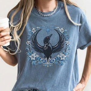 May include: Blue jean t-shirt featuring a detailed illustration of a black bird with outstretched wings, surrounded by a blue floral wreath and a star. The bird has a red eye. The shirt is a faded blue color.